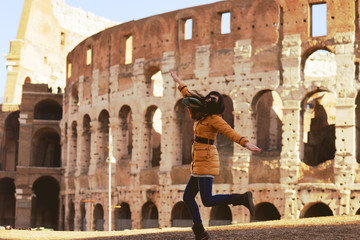 Young woman in winter clothes having fun on the background of the Winter Colosseum in Rome. The girl happily bounces on the photo of the Colosseum. Celebration of the New Year in Rome
