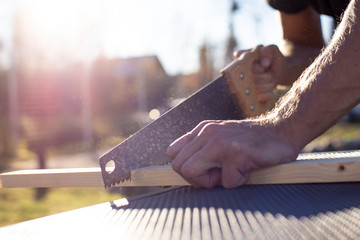 A man cuts a wooden product with a carpentry hand saw, in the sunshine on a warm summer day,...
