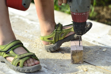 Little boy in sandals drills a hole in a piece of wood