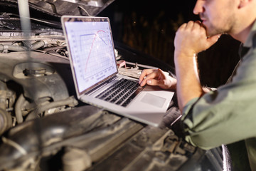 Mechanic in uniform looking intently at the laptop is connected to the motor vehicle for testing and adjustment. Mechanic carries out maintenance of the car. The removal of bugs of the engine.