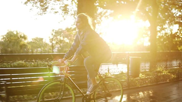 Pretty caucasian female riding a bicycle in the morning park or boulevard. Side view of a young woman riding a trekking bike, wearing sneakers and casual clothes