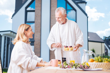 Fototapeta na wymiar Caring husband. Caring bearded husband bringing some fruit pancakes his appealing wife while having breakfast on summer terrace