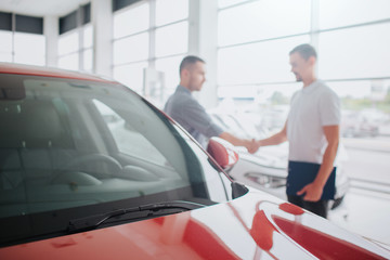 Customer and seller stand behind red car and shake hands. They have an agreement. Guy in white shirt holds tablet. They are inside in car store.
