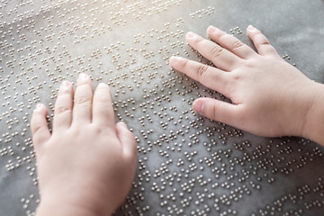 The blind kid's hand and fingers touching the Braille letters on the metal plate to understand an...