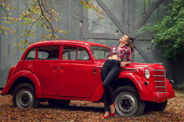 Pin-up girl in jeans and a plaid shirt is leaning on a russian red retro car. She smiles, basking in the sun.