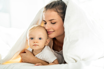 Portrait of young mother with infant baby girl liying on the bed covered with a white blanket