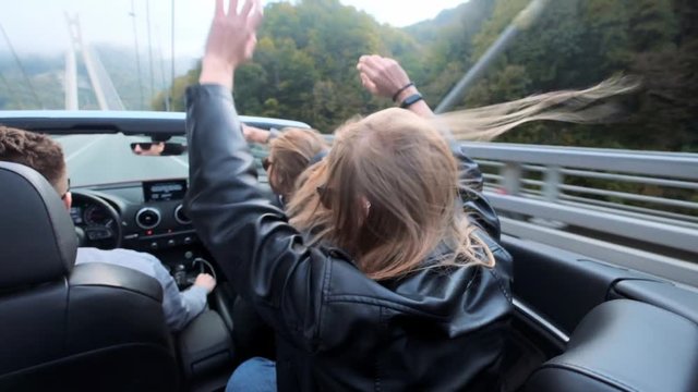 Two beautiful young girls ride in a red cabriolet among the mountains. Road on the highway. Dressed in black leather jackets. Hair fluttering in the wind. Enjoy travel and freedom. Slow motion 120fps
