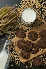 top view of the chocolate chip cookies on the wooden tray, a glass of soy milk, a pile of soybeans, a bottle of soy milk with dried wheat bouquet and the napery.