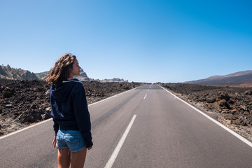 Naklejka premium Lonely beautiful young caucasian woman standing in front of a long way straight road in the middle of the lava landscape and mountains. travel and wanderlust experience and concept for lonely traveler