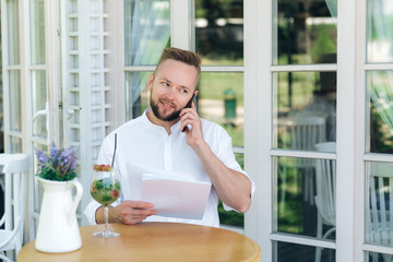 Close-up of a business man sitting at a table in a cafe, holding some important documents in his hand, talking on the phone with his personal assistant. The businessman has just signed an important