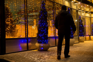 Man back to the viewer is on the night street, decorated with lights for Christmas.