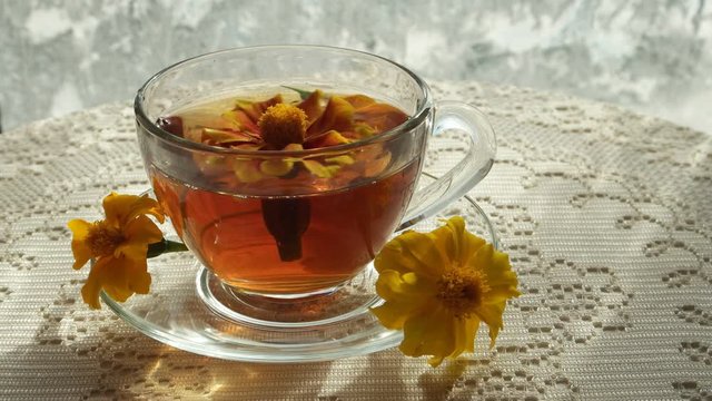 Marigold floats in a glass cup of herbal tea on the white table against ice flowers on the window glass in the sun, background