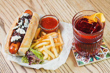 plate with hot dog with sausage and dor blue cheese, tomato sauce ketchup and french fried and old fashioned cocktail on the table in the bar