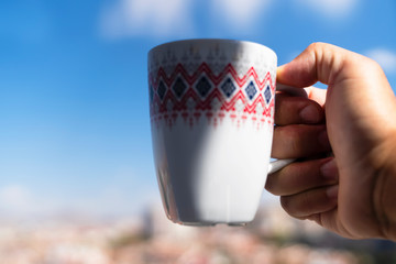 Human hand holding white and red decorative coffee cup up to the air with blue sky background with selective focus.