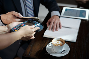 Businessman holding smartphone against terminal to pay for order in coffeeshop