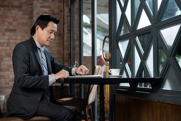 Mature Vietnamese business executive drinking coffee and reading news on tablet computer in...