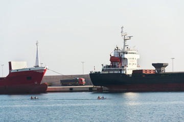 Freight truck stands between largo cargo ships in shipping harbor, oarsmen rowing in foreground sea, port Monopoli, Italy, freight digitization, transportation efficiency concept, sunny day copy space