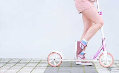 Girl in pink clothes rides on a pink scooter on the background of a white wall. Close-up photo of girl's legs, kick scooter and copyspace. White background