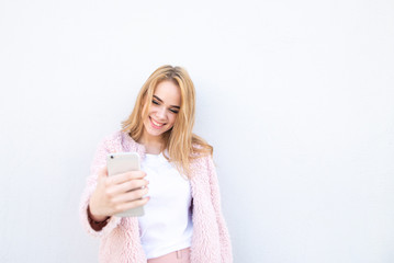Attractive girl wearing a white T-shirt and pink coat, smiles and uses a smartphone on a white background. Cute girl with a smartphone on the background of a white wall