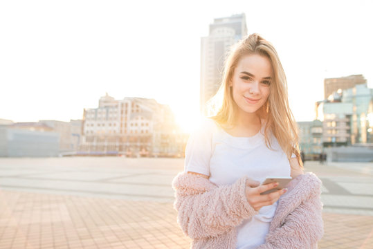 Smiling sweet girl standing with a smartphone in her hands, background of a city landscape in the sunset, looking into the camera and smiling.Street portrait of a cute girl with a smartphone.Copyspace