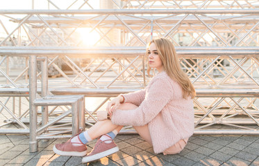 Stylish attractive girl in pink clothes sitting on the ground at the background of the sunset and...