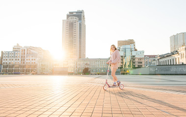 Pretty girl in pink clothes goes on a kick scooter on a beautiful city square in the sunset. Walk...