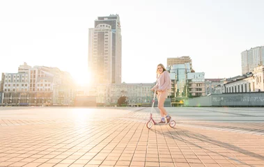 Foto op Canvas Pretty girl in pink clothes goes on a kick scooter on a beautiful city square in the sunset. Walk with a scooter on the sunset in the city. © bodnarphoto