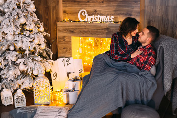 happy couple looking at each other sitting in a soft armchair and covered with a coverlet in a cozy room near the fireplace and christmas tree