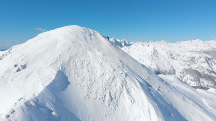 AERIAL: Unrecognizable skier hiking up the snowy mountain in sunny winter.
