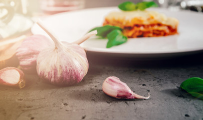 View of garlic. A piece of lasagne on a white plate on a dark stone counter and garlic. The concept of preparing lasagne and pasta.