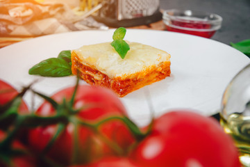View of lasagna. A piece of lasagne on a white plate on a dark stone counter and various ingredients like basil, cheese, oil, tomatoes. The concept of preparing lasagne and pasta.