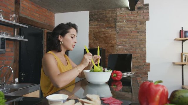 Wonderful young Caucasian woman with curly hair cooking tossed salad in the modern kitchen. Portrait of focused good-looking girl. Lifestyle.