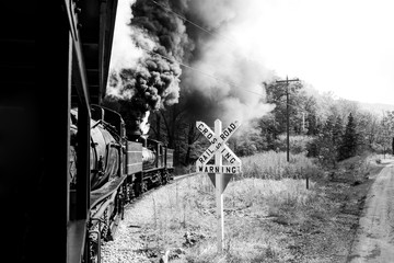 Steam locomotive rounding a bend at a railroad crossing with a giant plume of black smoke from it's coal burniong and white smoke from steam releasing