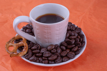 White cup of espresso with dark coffee, causer decorated coffee beans and pretzel on orange surface. Cheerfulness and positive mood concept.