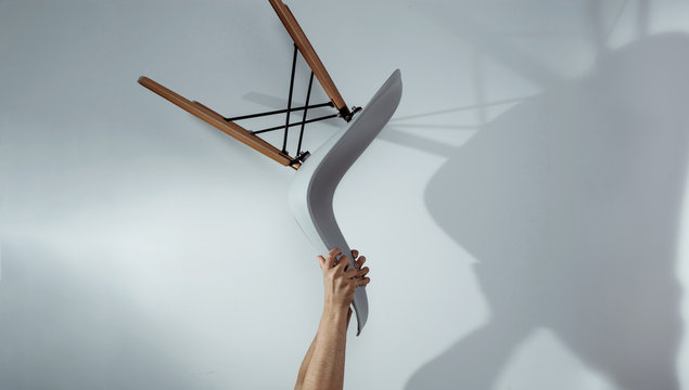 Hands Are Holding A Raised Chair On A Bright Gray Background. Man Throws A Chair, Devastates A Chair In The Office. The Concept Of Dissatisfaction With Work, From The Current Situation.