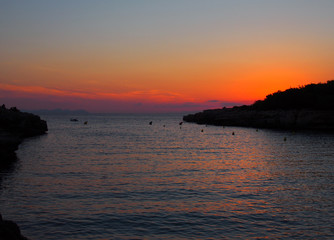 Naklejka premium glowing evening twilight after sunset in the cala santandria bay in ciutadela menorca with a beautiful orange sky reflected in a dark sea with the land in silhouette