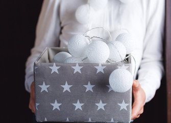 White cotton ball light garland in a grey basket on boys hands with stars sparkling at home. close up, bright lights, festive atmosphere. home decoration. New year and Christmas celebration concept