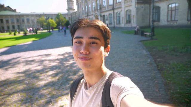 Happy confident asian guy is holding selfie camera and taking video in sunlights near beautiful old building, chilling