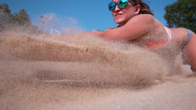 SLOW MOTION, CLOSE UP, DOF: Smiling Caucasian female volleyball player dives and strikes ball with her hands. Young woman on active summer vacation playing beach volleyball on a sunny day. Sand flying