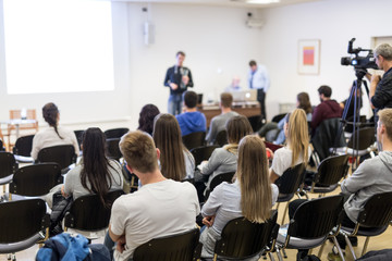 Speaker lecturing in lecture hall at university. Students listening to lecture and making notes.