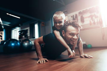 Fototapeta na wymiar Man Is Doing Push Ups In Gym With Son On A Back.