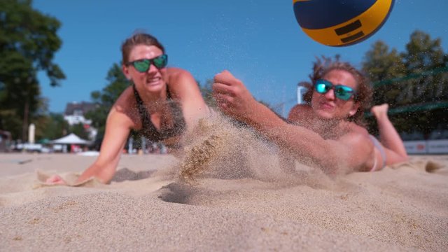 SLOW MOTION, CLOSE UP, DOF: Athletic women in bikinis jump towards the ball and crash in the hot sand while playing beach volleyball. Cheerful friends on summer vacation playing volleyball in the sun.