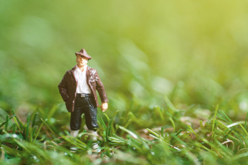 Selective focus miniature people wearing leather jacket and cowboy hat standing in green field with sun raise flare, cattleman.