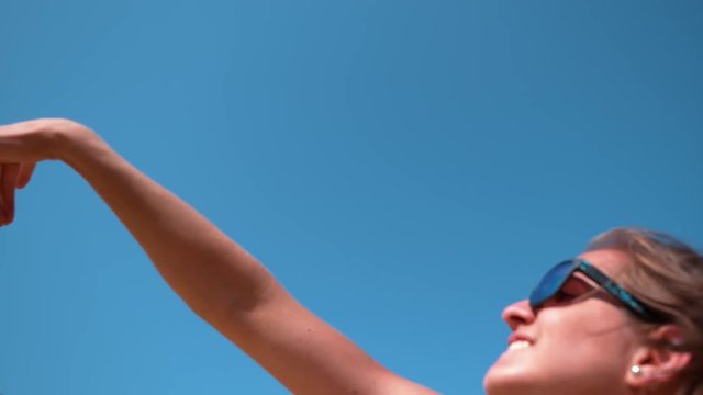SLOW MOTION, CLOSE UP, DOF: Beautiful young Caucasian woman playing volleyball strikes the ball above her head on a sunny day. Girl wearing blue sunglasses serves the ball during beach volleyball game