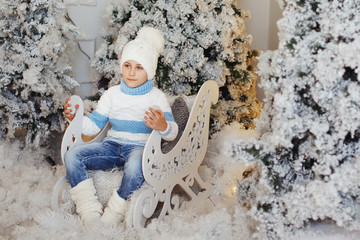 Naklejka premium Boy in a white and blue sweater and white hat is sitting in a decorative sleigh near the Christmas tree.