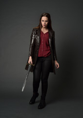 Fototapeta premium full length portrait of brunette girl wearing long leather coat and boots. standing pose and holding a sword on grey studio background.