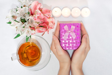 Dressing table with flowers, a cup of tea, candles and women's hands holding a gift box.