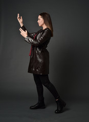 full length portrait of brunette girl wearing long leather coat and boots. standing pose  on grey studio background.