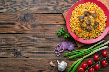 Red plate with pilaf on a brown wooden table. On the table is red pepper, green onions, garlic, cherry tomatoes, red napkin, spoon. Top view. Flat lay.