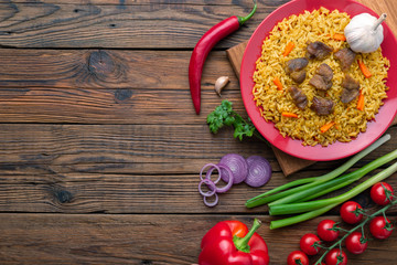 Red plate with pilaf on a brown wooden table. On the table is red pepper, green onions, garlic, cherry tomatoes, red napkin, spoon. Top view. Flat lay.